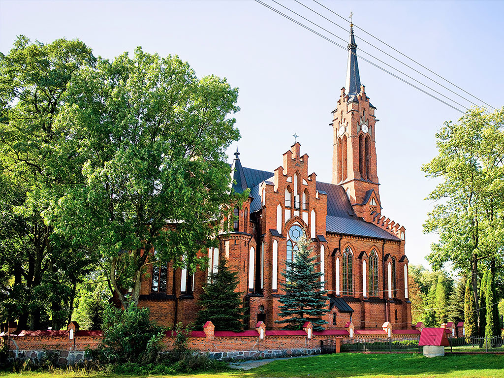 Kościół z cegły w otoczeniu drzew i ogrodzony parkanem w tle niebieskie niebo.