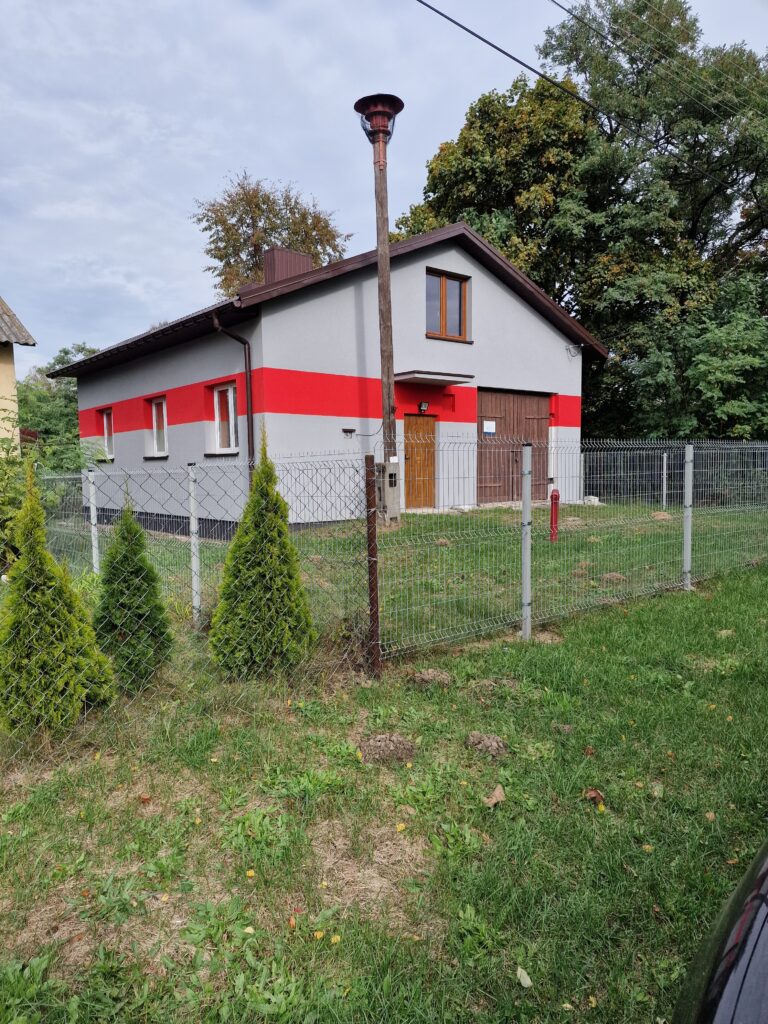 Na obrazku widoczny jest dom, który ma czerwone i białe kolory. Wygląda na nowoczesny oraz dobrze utrzymany, a jego białe elementy kontrastują z szeroką elewacją.