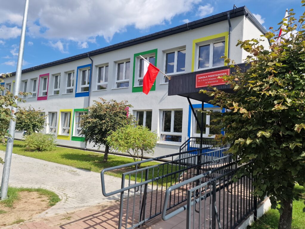 Na obrazku widocznym jest budynek szkoły z kolorowymi oknami. Zwieńczeniem konstrukcji jest maszt, na czele powiewa flaga.