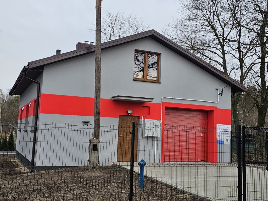 Obrazek przedstawia budynek wykończony w kolorze czerwonym i szarym. Wokół niego znajduje się ogrodzenie.