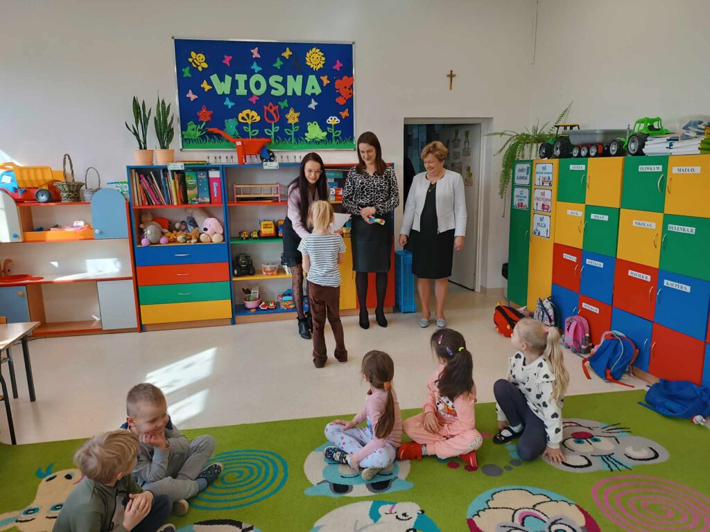 Na obrazku widać grupę dzieci siedzących na dywanie w klasie, gdy jest ona dostępna w pobliżu. W tle znajduje się półka z zabawkami i przegródkami.