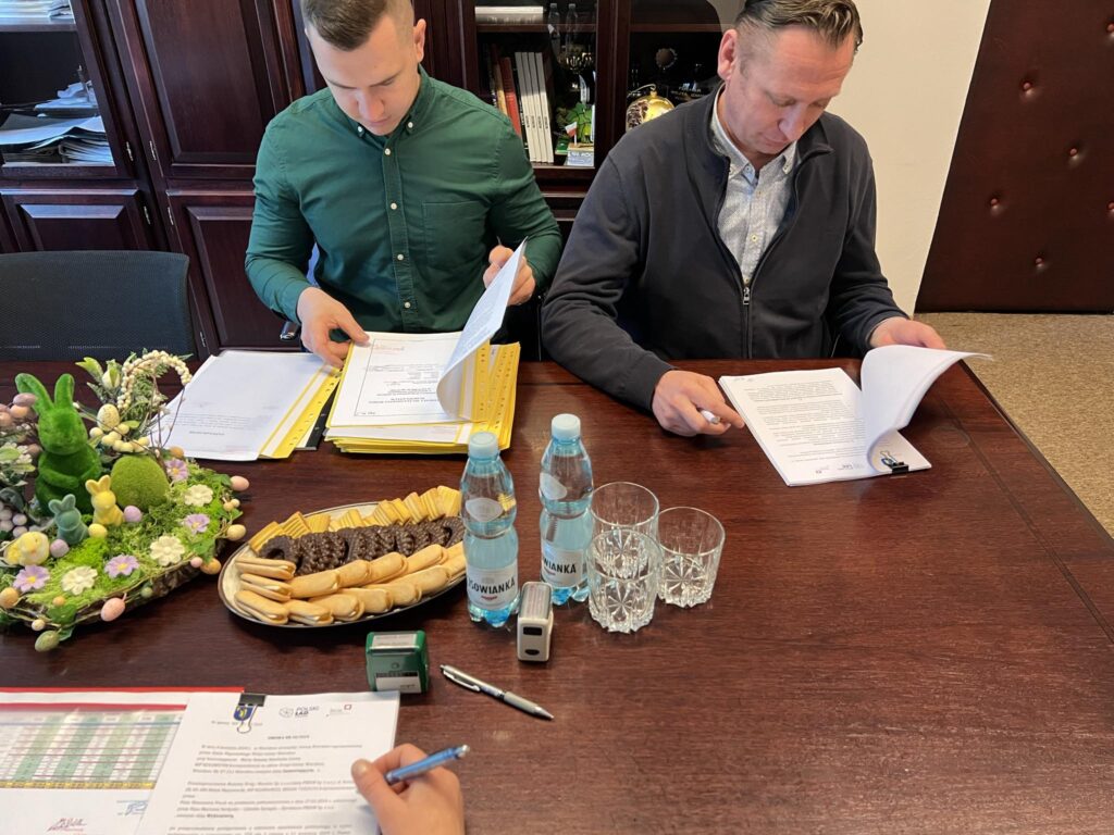 Na obrazku dwóch mężczyzn przeglądających dokumenty siedzące przy stole konferencyjnym. Na stole znajdują się również lekkie opakowania i opakowania, o charakterze nieformalnym.
