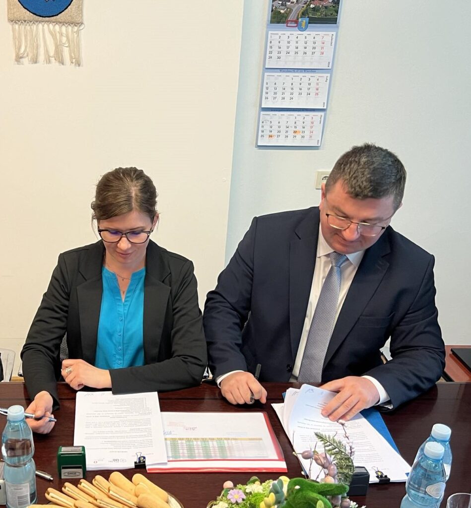 Na obrazku widać dwie osoby siedzące przy biurku i podpisujące dokumenty. Oboje skupieni są na czytaniu i formalizowaniu tych papierów.