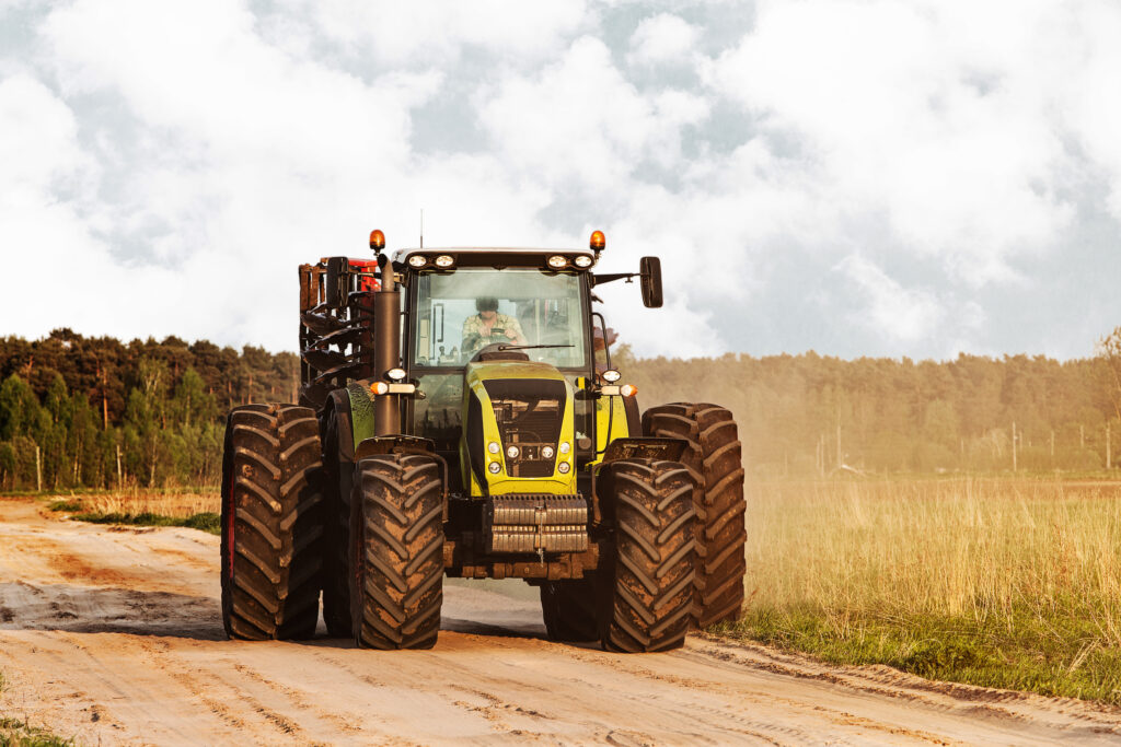 Najprawdopodobniej pojawi się duży traktor z końcowymi oponami, jadący polnej drodze na wiejskim. Otaczają go trawa i drzewo, a nad nim unosi się zachmurzone niebo.