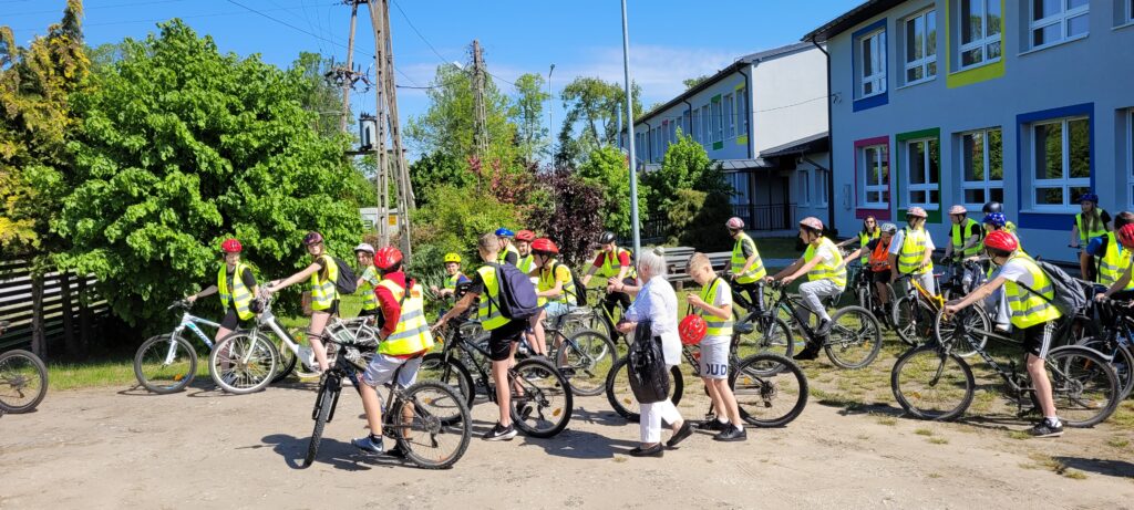 Grupa rowerzystów w kamizelkach odblaskowych i kaskach, na tle zieleni i budynku z kolorowymi ramami okien.