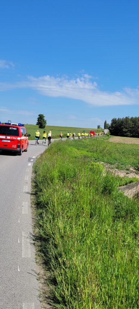 Czerwony wóz strażacki jedzie obok grupy rowerzystów w żółtych kamizelkach. Droga otoczona jest zielonymi polami. Na niebie widać białe chmury.