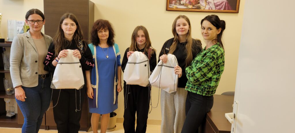 Sześć osób stoi w pomieszczeniu o jasnych ścianach. Trzy trzymają białe worki. W tle ciemne półki i obraz.
