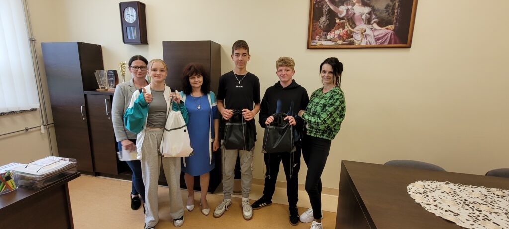 Na zdjęciu widocznych jest sześć osób w pomieszczeniu biurowym. W tle widać szafki, zegar i obraz na jasnej ścianie. Osoby trzymają torby, ubrane są w casualowe stroje.