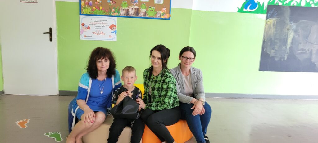 Na zdjęciu widać trzy kobiety i dziecko na tle zielonej ściany z dekoracjami. Kobiety siedzą na kolorowych pufach. W tle tablica z rysunkami żabek.