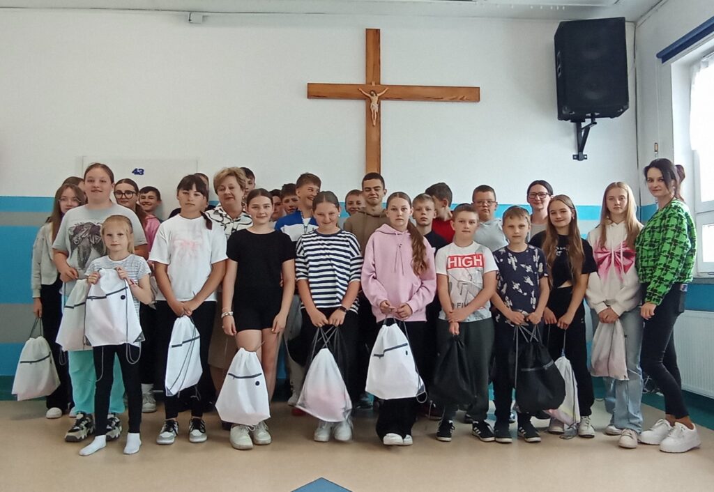 Grupa dzieci i dorosły stoją w jasnym pomieszczeniu z krzyżem na ścianie. Trzymają białe torby, a ściany są biało-niebieskie. W tle znajduje się duży głośnik i okno.