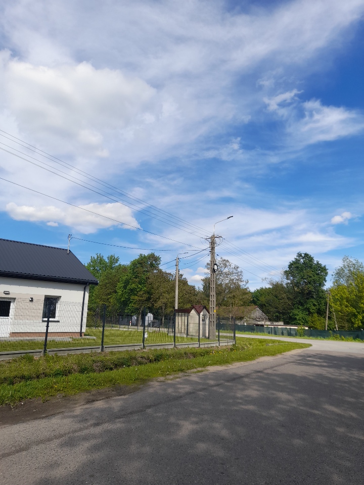 Biały budynek z ciemnym dachem obok drogi, ogrodzony płotem. Niebo jest błękitne z białymi chmurami, w tle drzewa. Latarnia i linie energetyczne widoczne.