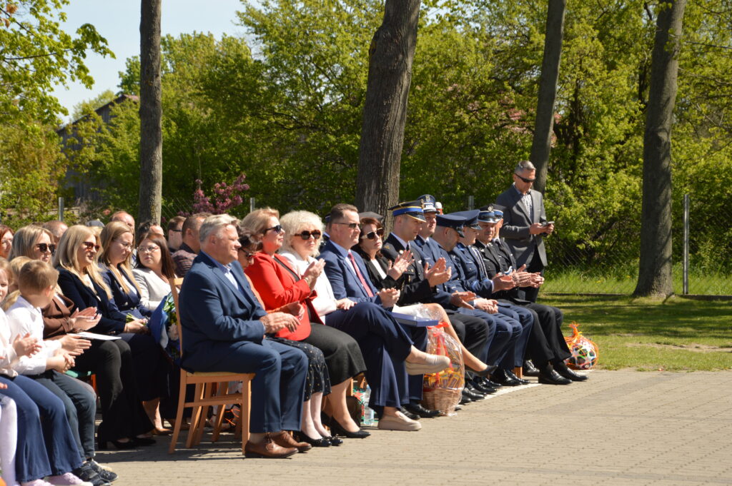 Ludzie siedzą na zewnątrz w parku, klaskając; większość w formalnych ubraniach. Tło to zielone drzewa i jasne niebo. Po prawej widoczni oficjele w mundurach.