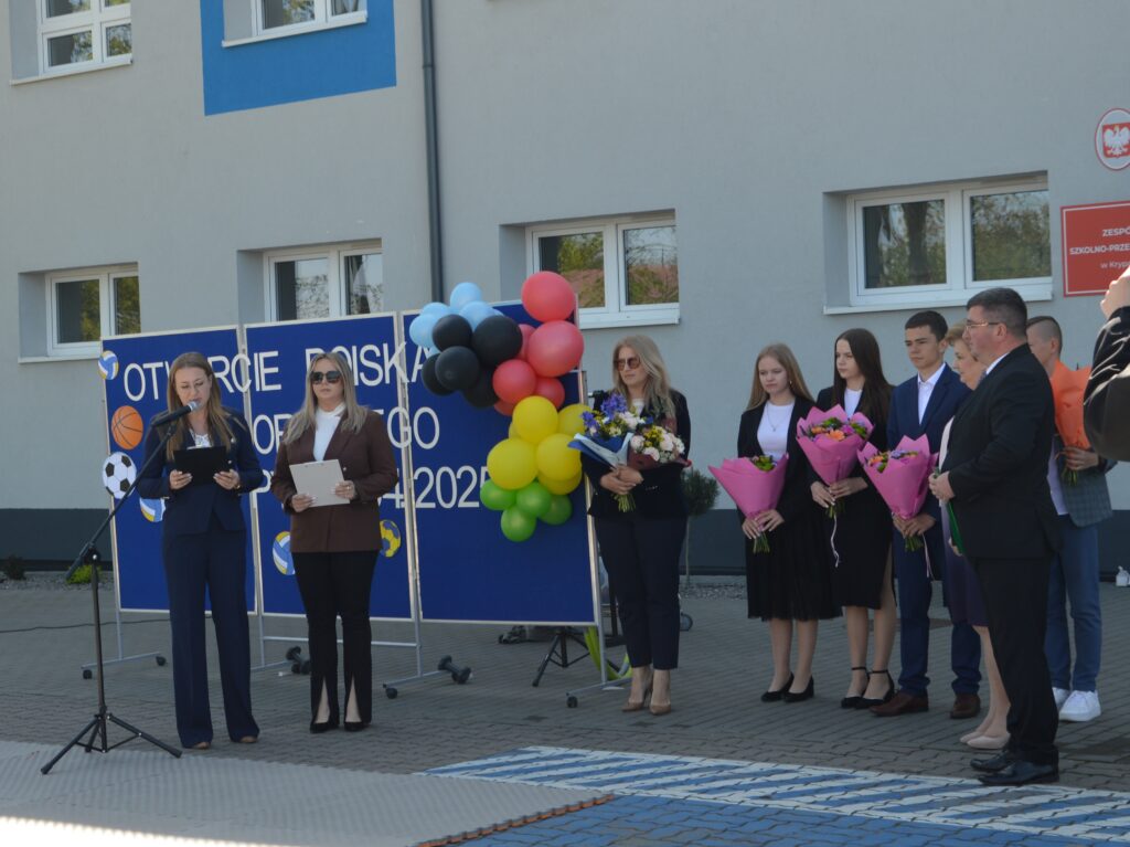 Ceremonia otwarcia boiska, osoby z kwiatami na tle budynku. Tablica z balonami i napisem, kolory jasne.