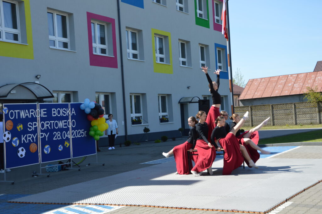 Grupa tancerek w czerwonych spódnicach przed budynkiem z kolorowymi ramkami okien. Scena na świeżym powietrzu. Tło z balonami i plakatem otwarcia boiska.