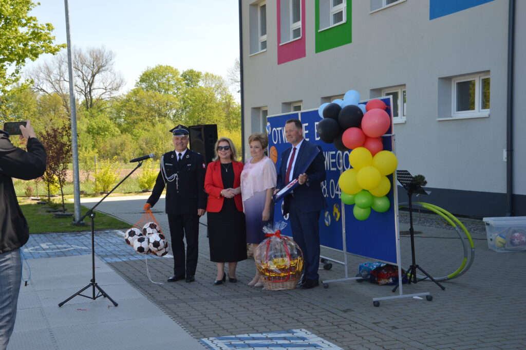 Grupa osób przy mikrofonie na tle budynku z kolorowymi balonami i tablicą. Słoneczny dzień, zieleń w tle.