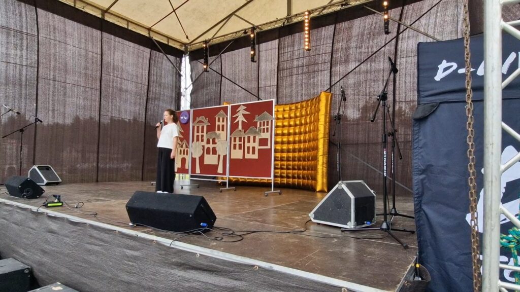 Scena z dziewczynką trzymającą mikrofon. W tle dekoracje w kolorze bordowo-złotym przedstawiające budynki. Jasne oświetlenie, mikrofony na statywach po bokach.