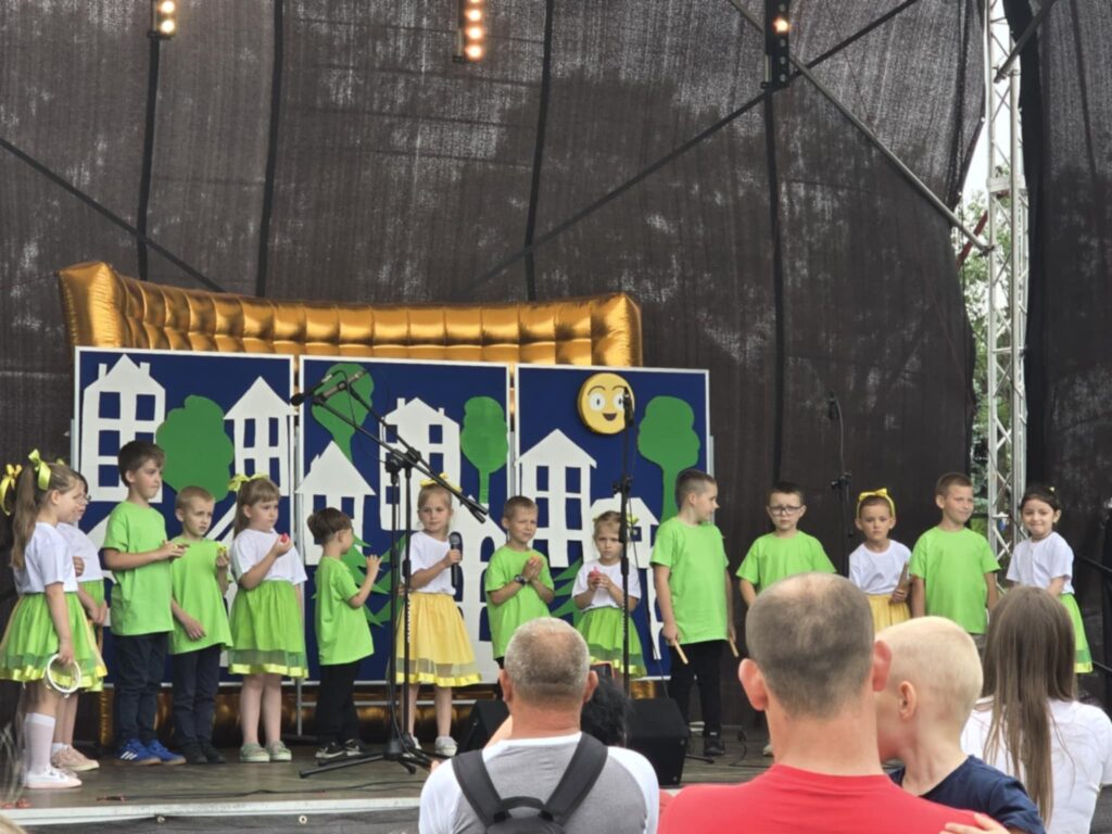 Dzieci w zielonych i żółtych strojach występują na scenie. Tło to rysunki białych domów na niebieskim tle. Publiczność ogląda z dołu sceny.