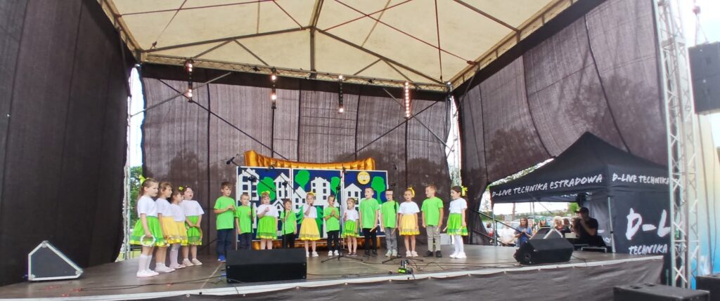 Grupa dzieci w kolorowych strojach występuje na scenie. Tło z rysunkami domków i słońca. Scena zadaszona, czarne kurtyny.
