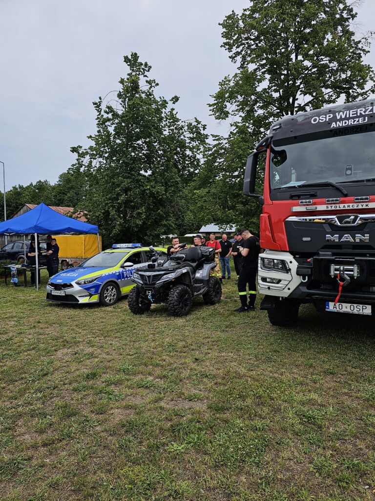Na trawie stoi czerwony wóz strażacki, quad i niebieski samochód policyjny. W tle widoczny jest niebiesko-żółty namiot. Dookoła są drzewa i kilka osób w mundurach.