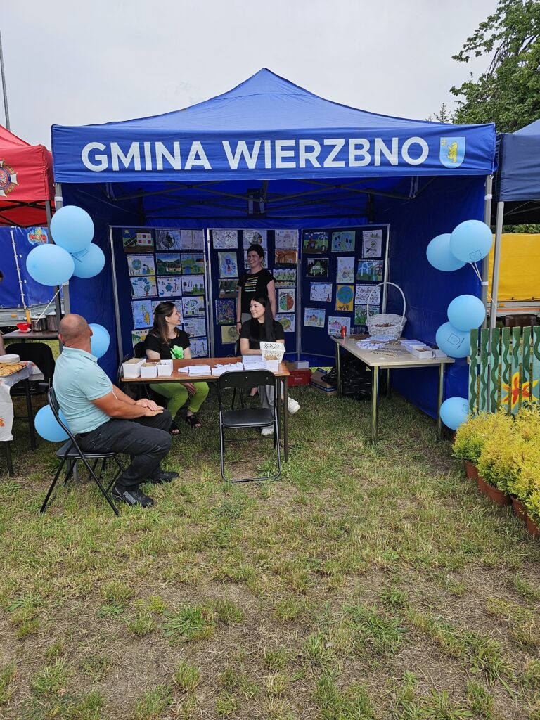 Niebieski namiot z napisem Gmina Wierzbno stoi na trawie. Wewnątrz rozstawione kolorowe rysunki na ścianie. Trzy osoby siedzą przy stoliku z balonami.