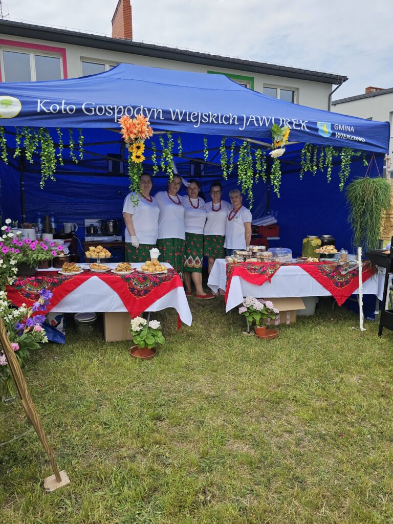 Niebieski namiot z napisem, w którym pięć osób w ludowych strojach stoi przy stołach z regionalnymi potrawami. 앞에는 czerwone obrusy i zielone rośliny. Tło stanowi budynek z oknami pod jasnym niebem.