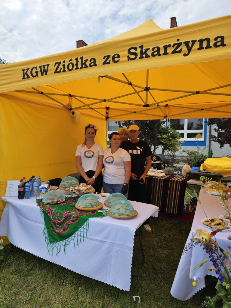 Namiot żółty z napisem, trzy osoby, stół z ciastami pod niebieskimi pokrywkami, zielona serweta, kwiaty na białym obrusie.
