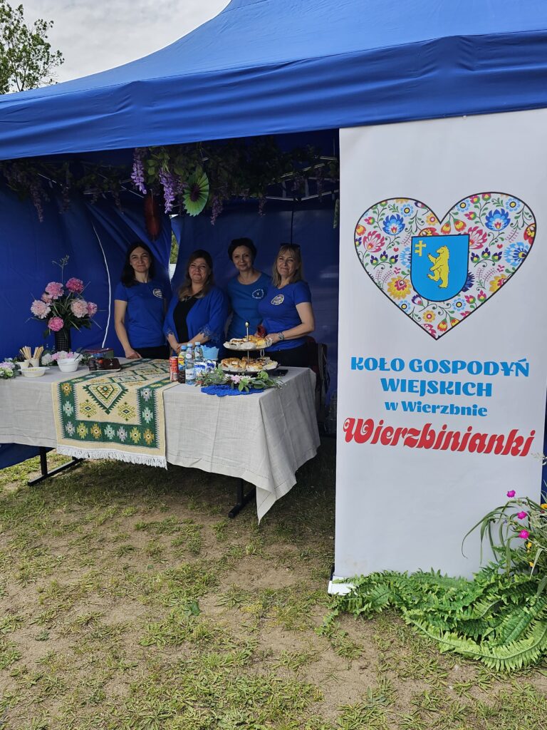 Kobiety w niebieskich koszulkach stoją pod niebieskim namiotem. Stół z białym obrusem ozdobiony jest potrawami i kwiatami. Obok znajduje się tablica z napisem Koło Gospodyń Wiejskich Wierzbinanki.