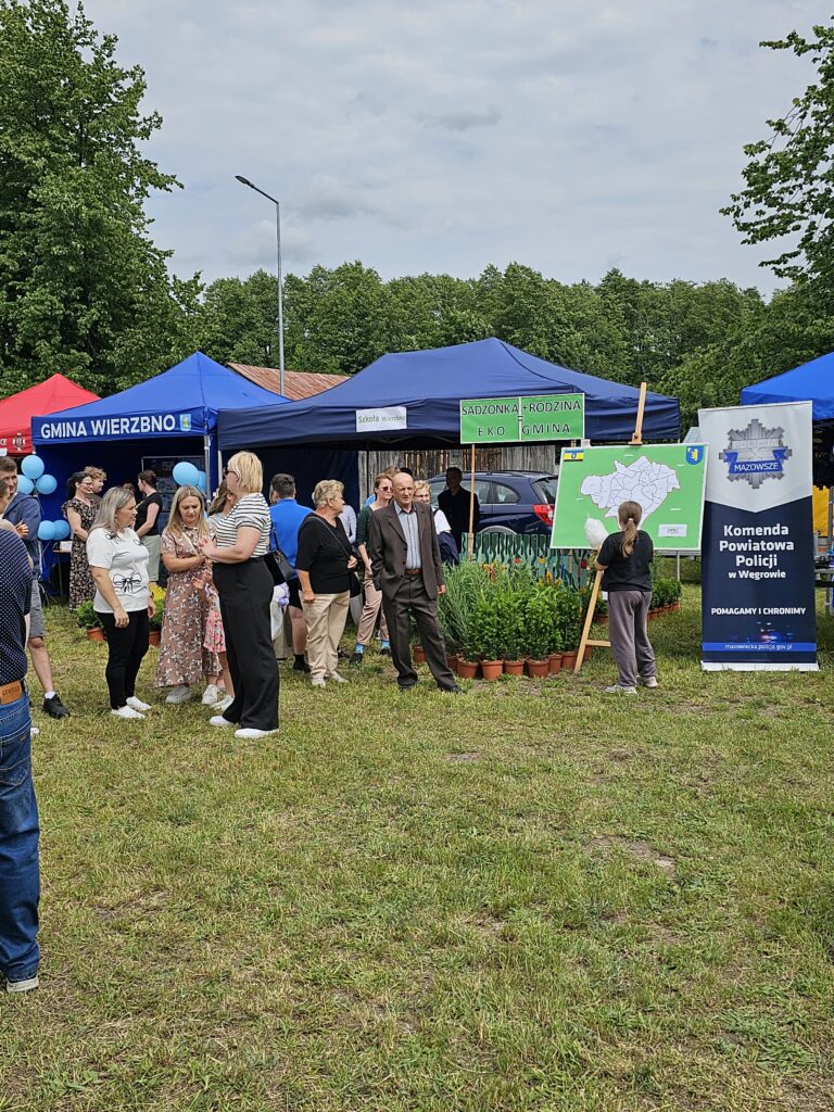 Na zdjęciu widoczni są ludzie przy stoiskach z niebieskimi i czerwonymi namiotami na tle drzew. W centrum mapa z napisem Komenda Powiatowa Policji. Trawa i rośliny w doniczkach dodają zieleni.