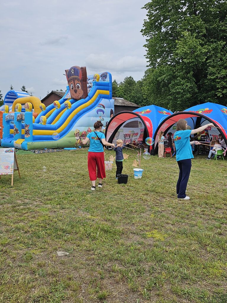 Na tle zielonego parku, widoczna jest wielka niebiesko-żółta zjeżdżalnia z postaciami z bajki. Dwoje ludzi w niebieskich koszulkach bawi się bańkami mydlanymi. Obok ustawiono kolorowe namioty z logotypami