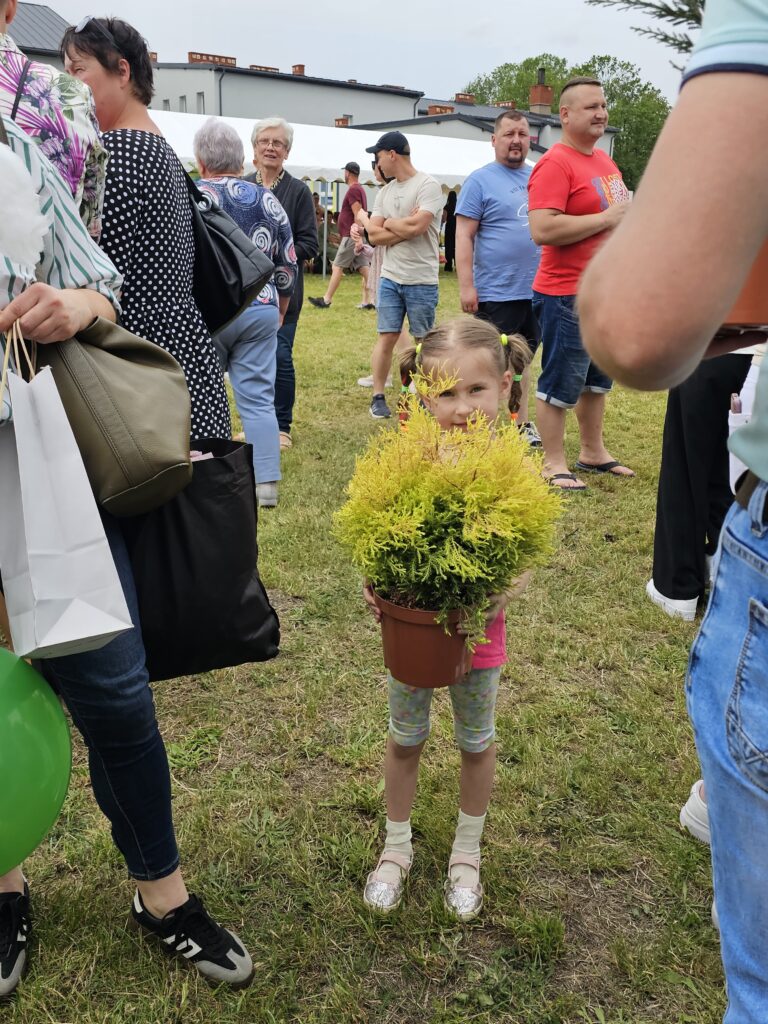 Na trawie stoi dziewczynka z doniczką żółtych kwiatów. W tle znajdują się ludzie i białe namioty. Dzień jest pochmurny.