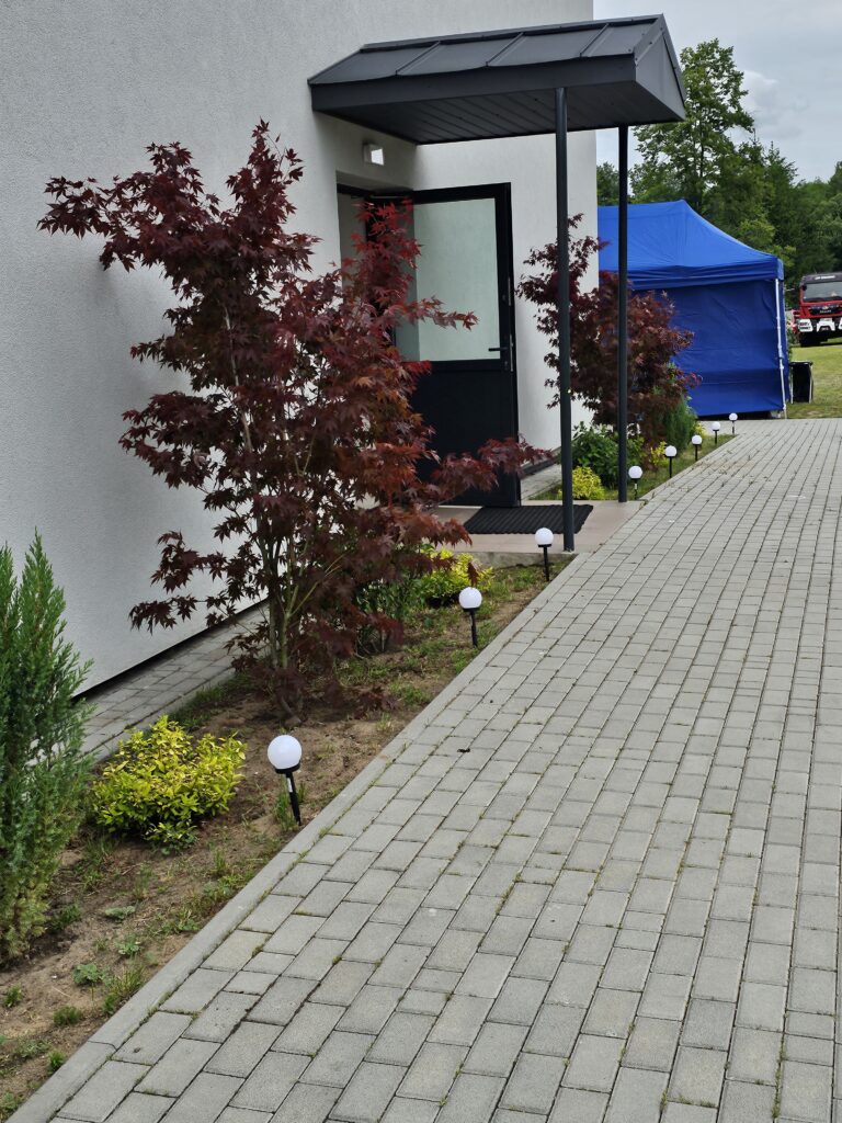 Ścieżka z szarej kostki brukowej prowadzi do drzwi z daszkiem, otoczona czerwonymi krzewami i oświetleniem. W tle niebieski namiot.