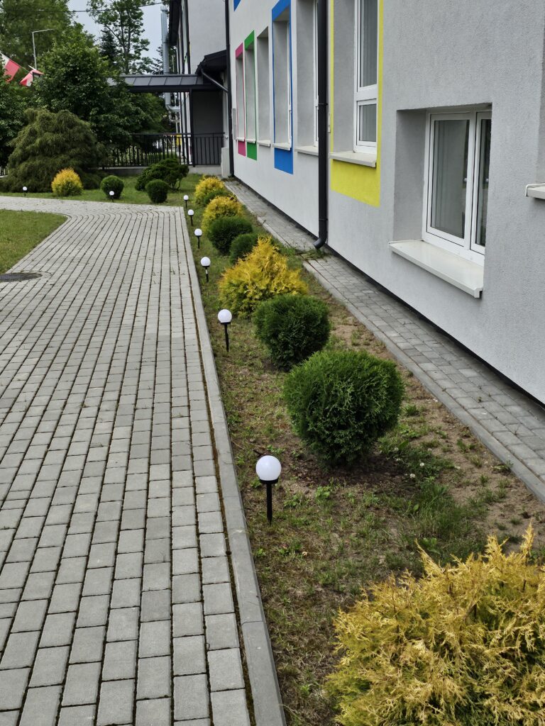 Budynek z szarymi ścianami i kolorowymi obramowaniami okien. Przed nim biegnie wyłożona kostką ścieżka z zielonymi krzewami. Latarnie ogrodowe dodają elegancji.