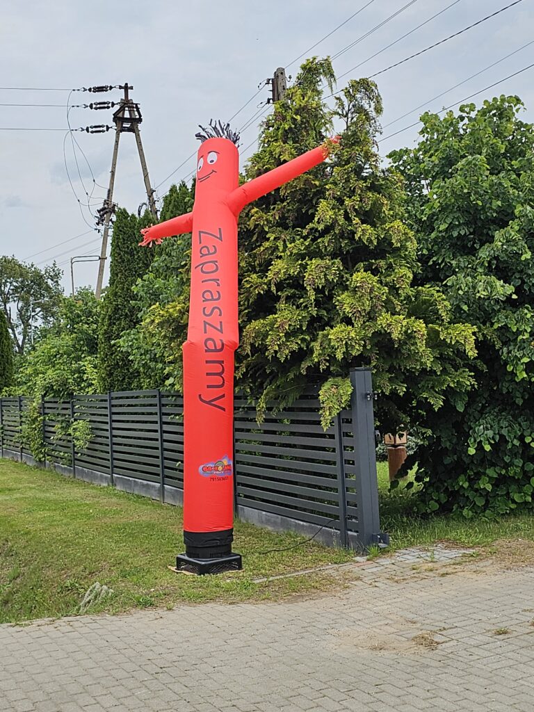 Czerwony nadmuchiwany ludzik z napisem Zapraszamy stoi przed zielonym żywopłotem. W tle widać szary płot i słup elektryczny. Trawnik otacza chodnik.