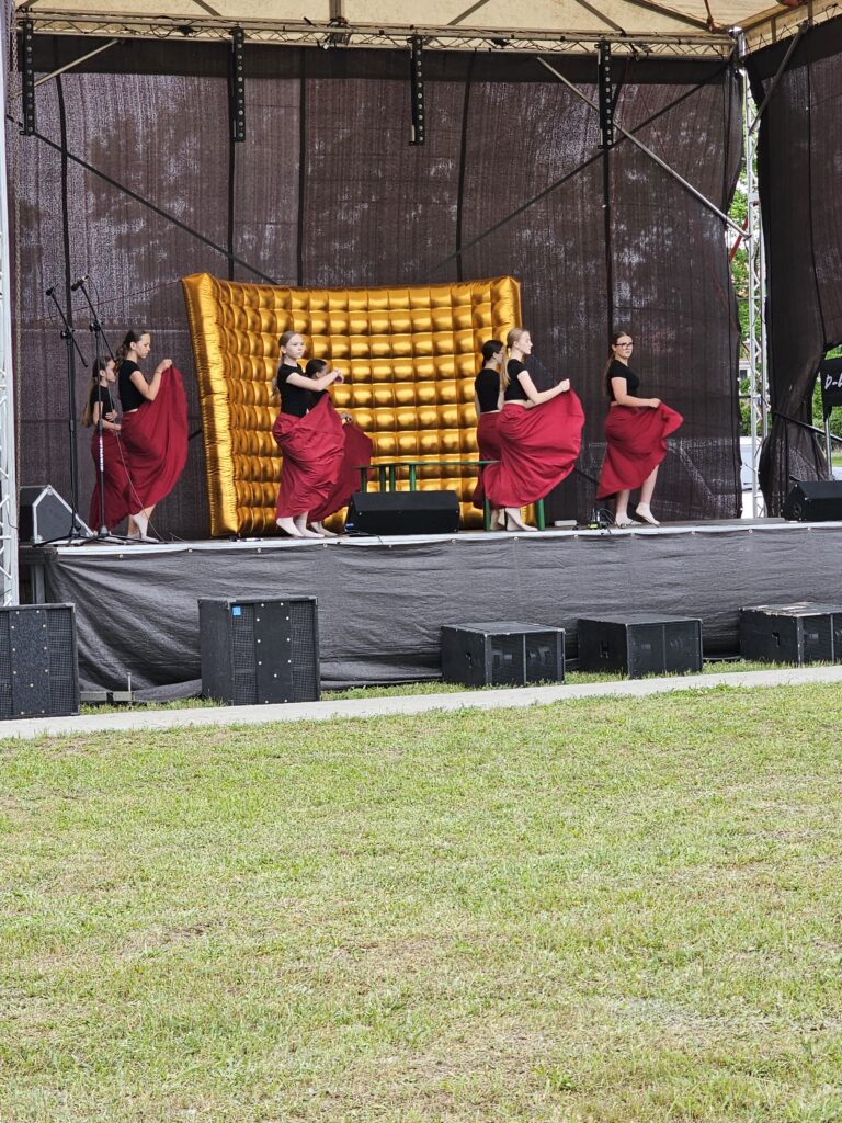 Młode tancerki w czarnych koszulkach i czerwonych spódnicach występują na scenie. Tło stanowi złoty, pikowany panel. Scena znajduje się na trawie, z profesjonalnym nagłośnieniem.