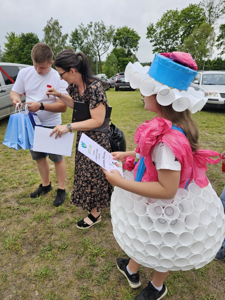 Trzy osoby stoją na trawniku. Jedna z nich jest w kostiumie z białych kubków, niebieskim kapeluszu i różowym szalu. W tle są zaparkowane auta i drzewa.