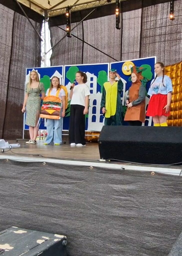Grupa dzieci na scenie w kolorowych kostiumach. Tło przedstawia niebieskie i zielone dekoracje. Chłodne światło sceniczne.