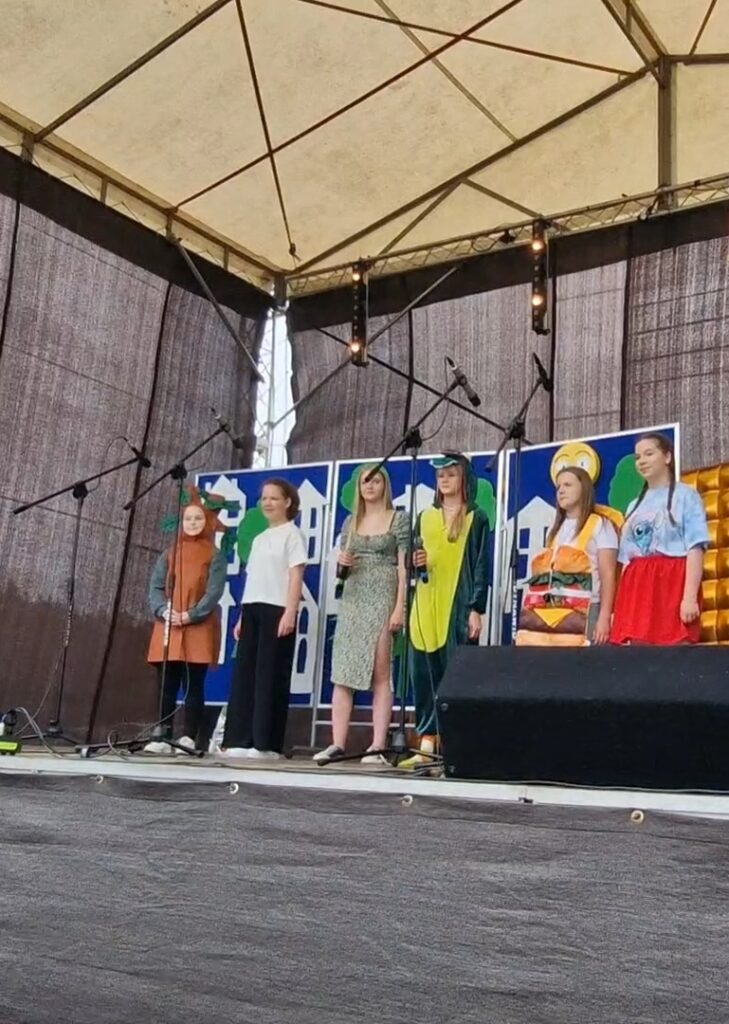 Na scenie pod namiotem, sześć osób w kolorowych kostiumach. Tło z niebieskimi i białymi wzorami. Mikrofony ustawione przed nimi.