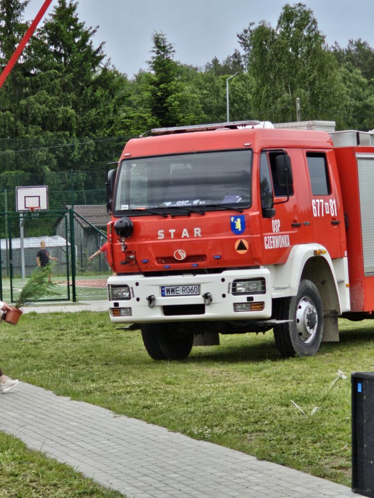 Czerwony wóz strażacki stoi na trawie obok boiska sportowego. W tle są zielone drzewa i siatka. Pogoda jest pochmurna.