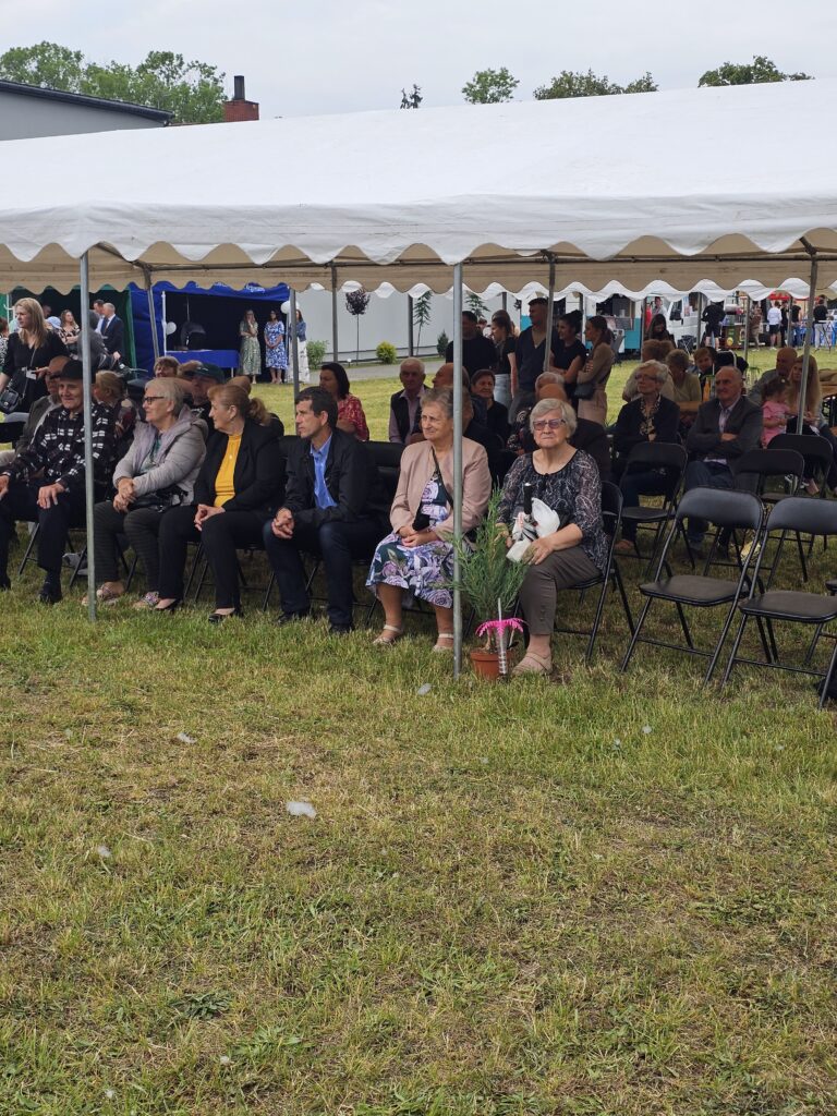 Grupa ludzi siedzi pod białym namiotem na zewnątrz. Przed nimi zielona trawa, a w tle są drzewa i budynek. Uczestnicy są elegancko ubrani.