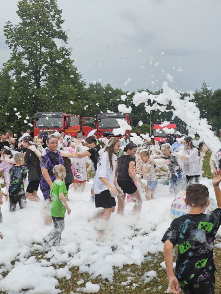 Na zdjęciu jest grupa ludzi bawiących się w pianie na trawniku. W tle stoją dwa czerwone wozy strażackie, a niebo jest pochmurne. Dominują kolory bieli piany oraz zieleń trawy i drzew.