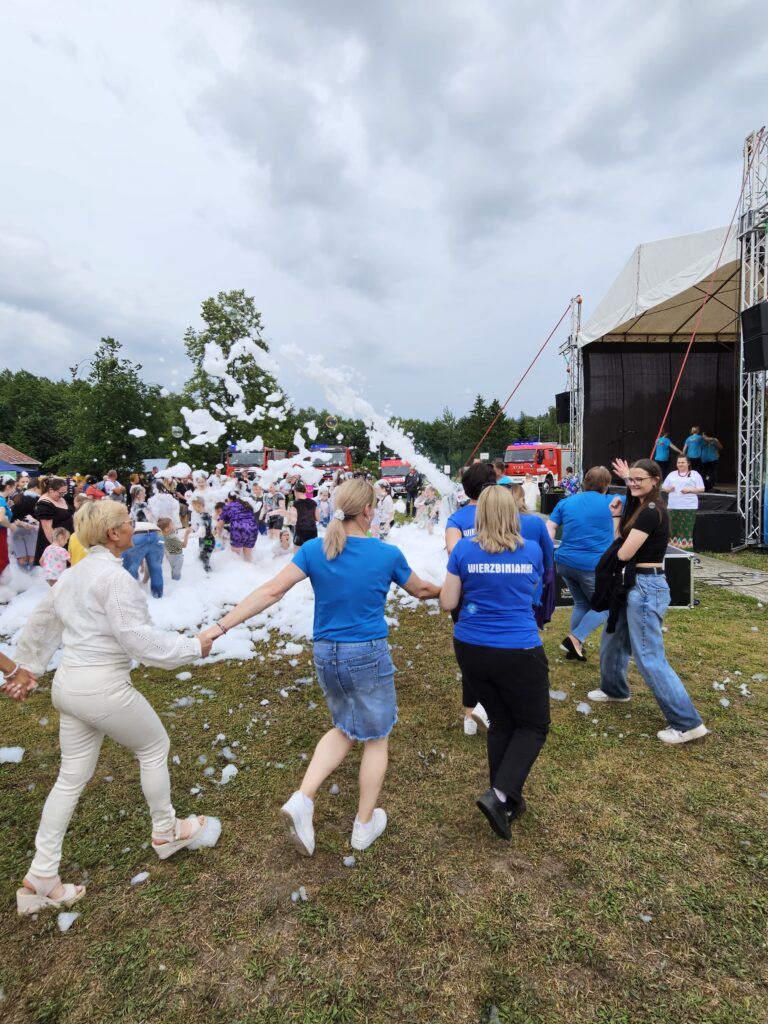 Grupa ludzi bawi się w pianie na trawiastej imprezie plenerowej. Większość ubrana na niebiesko, z tyłu scena i drzewa. Na niebie są chmury, a wokół wiele osób.