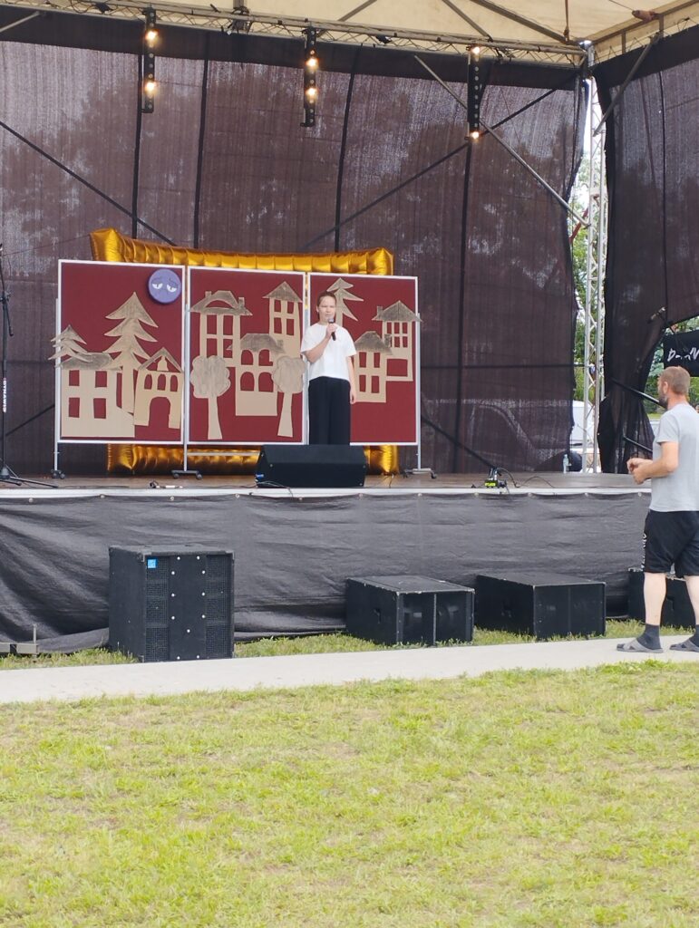 Na scenie występuje osoba przy mikrofonie, z tłem z czerwonymi panelami ozdobionymi złotymi budynkami. Tło jest ciemne, scena ustawiona na zewnątrz. Przed sceną trawa, po prawej stronie stoi inna osoba.