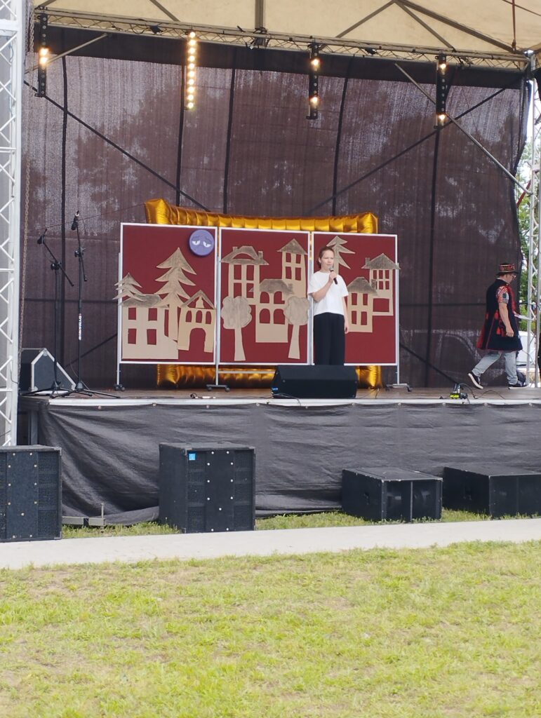 Scena plenerowa z brązowym tłem i dekoracjami w kształcie domów. Przed dekoracją stoi osoba w białej koszulce, obok stoi inna osoba. Tło sceniczne ma żółty przebłysk, a na trawie leżą głośniki.
