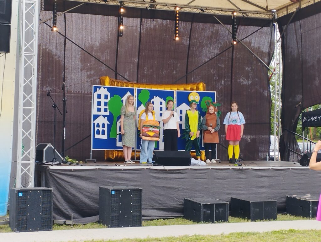 Na scenie stoi sześć osób, przebranych kolorowo. Tło zdobią niebiesko-zielone dekoracje. Scena na świeżym powietrzu.