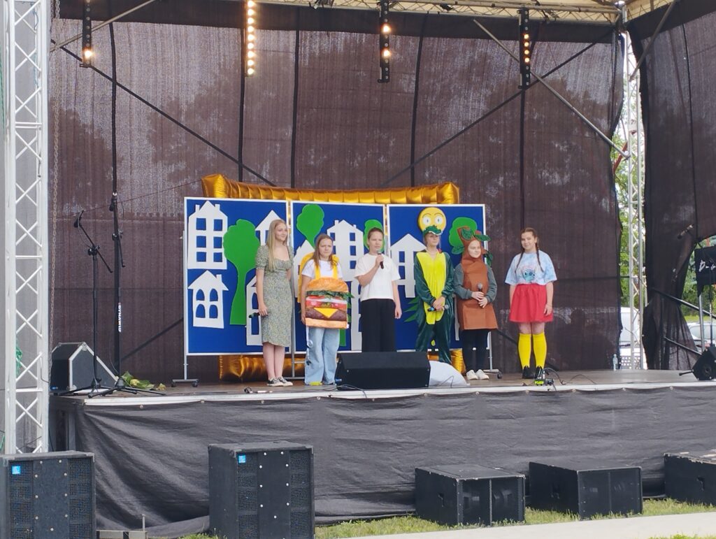 Sześć osób stoi na scenie przed kolorowym tłem z domami i drzewami. Występują w kostiumach, trzymają mikrofony. Scena otoczona jest czarnymi kurtynami.