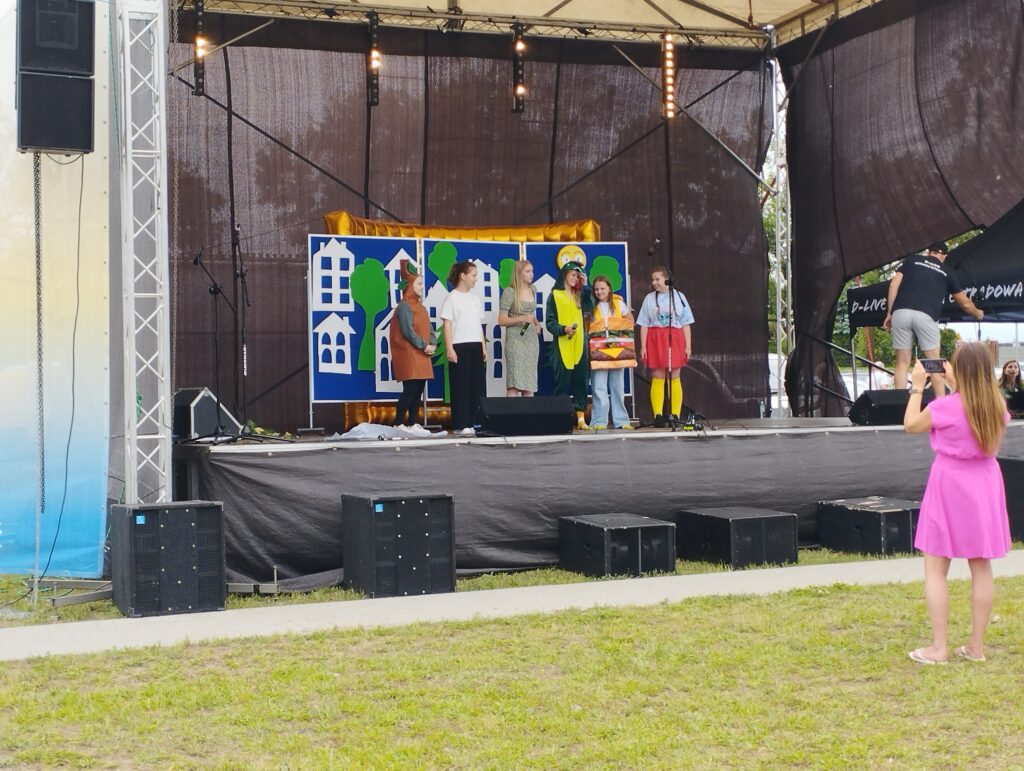 Grupa osób w kolorowych strojach występuje na scenie. W tle dekoracje z białymi sylwetkami domów. Na pierwszym planie osoba w różowej sukience fotografuje scenę.
