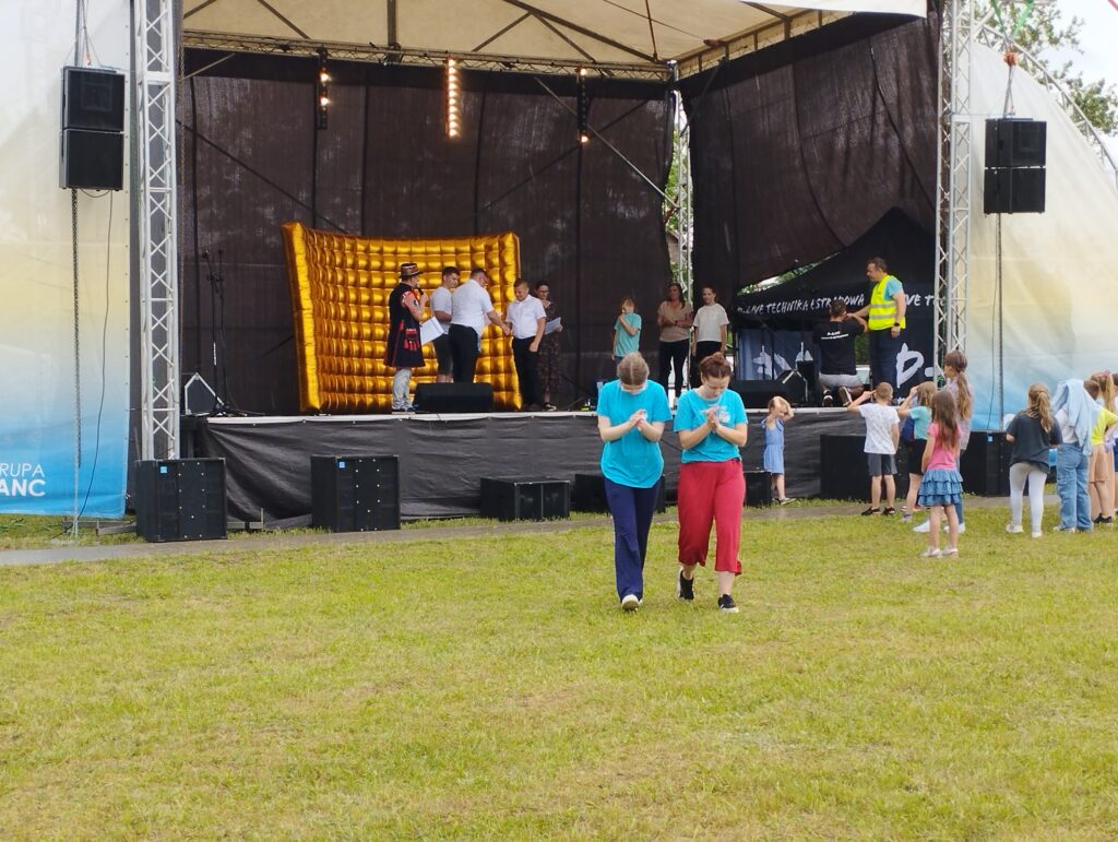 Na trawiastej scenie plenerowej grupa ludzi występuje przed publicznością. Niektórzy uczestnicy w niebieskich koszulkach tańczą. W tle widoczny jest złoty element dekoracyjny.