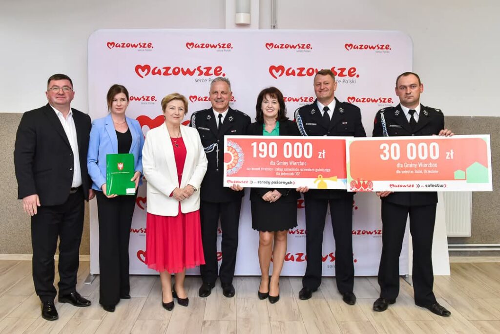 Grupa osób stoi, trzymając duże czeki; tło z logo Mazowsze. Dominują barwy czerwone, czarne i białe. Uśmiechnięta atmosfera.