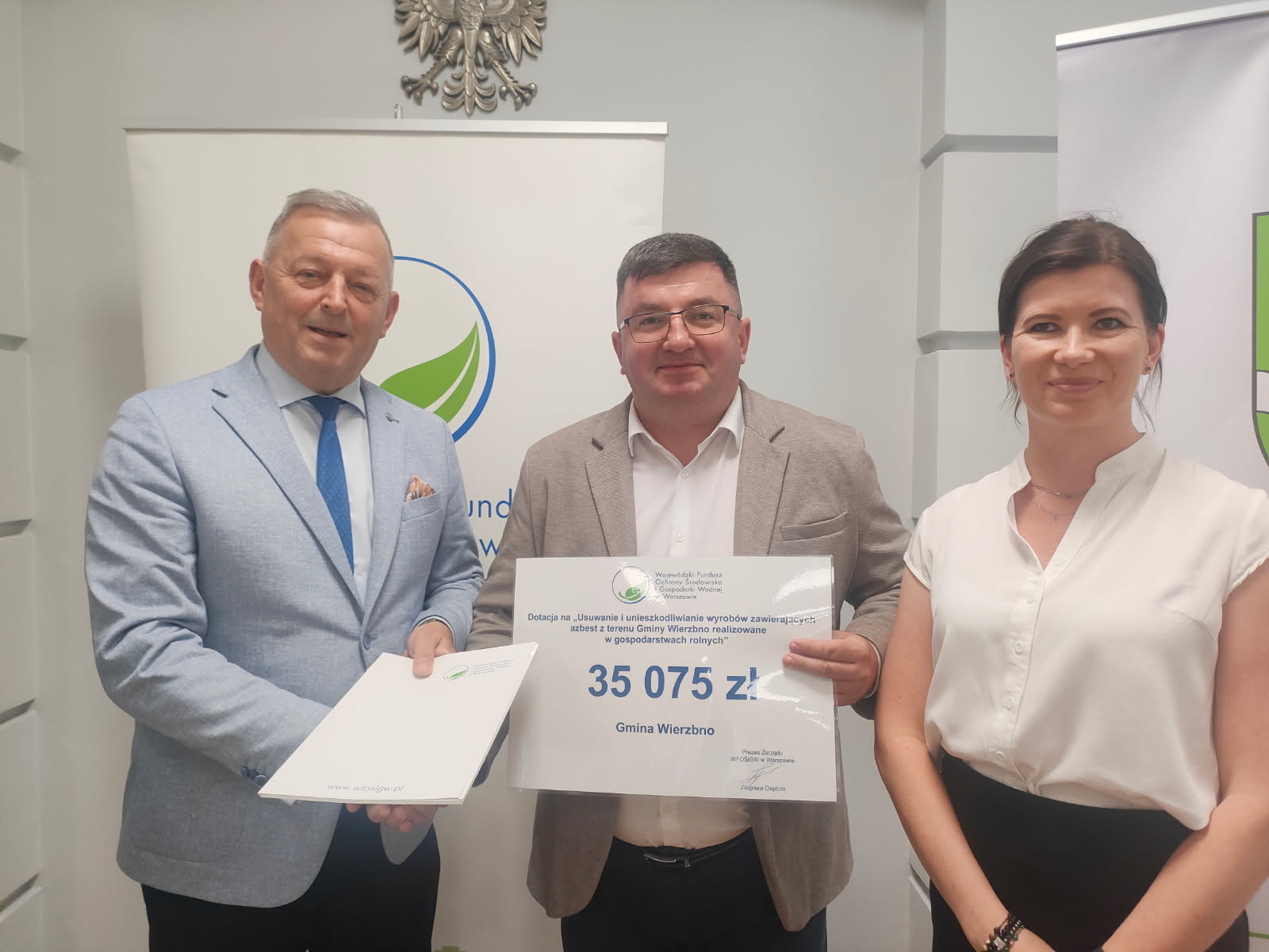 Trzy osoby stoją w jasnym pokoju, trzymając tabliczkę z kwotą 35 075 zł. Mężczyzna z lewej ma jasny garnitur, kobieta po prawej jest ubrana na biało. W tle znajduje się biały baner z zielonym logo.