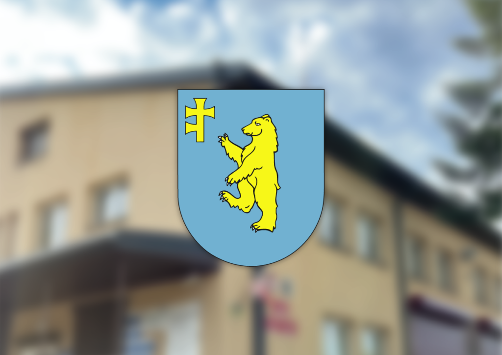 Herb na niebieskim tle przedstawia żółtego niedźwiedzia i krzyż. W tle rozmyty budynek w kolorze beżowym. Na niebie białe chmury.